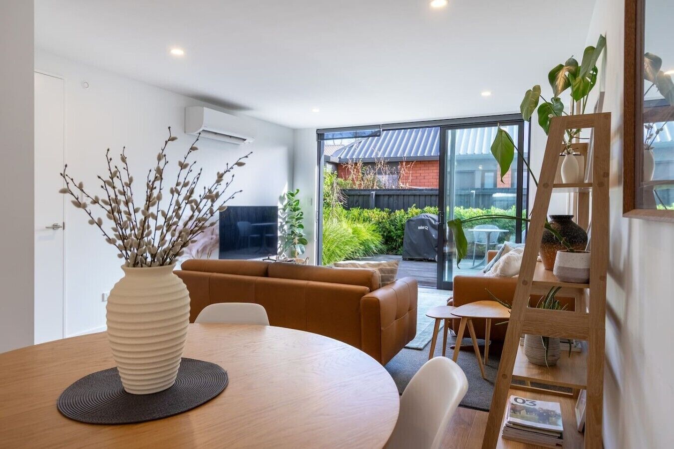 A modern living room with a sofa, a dining table, and a coffee table. There is a large window providing a view of the outdoors.