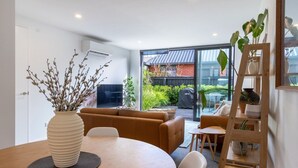 Ein modernes Wohnzimmer mit einer Couch, einem Esstisch und einem Couchtisch. Ein großes Fenster bietet Blick ins Freie.