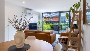 A modern living room with a sofa, a dining table, and a coffee table. There is a large window providing a view of the outdoors.