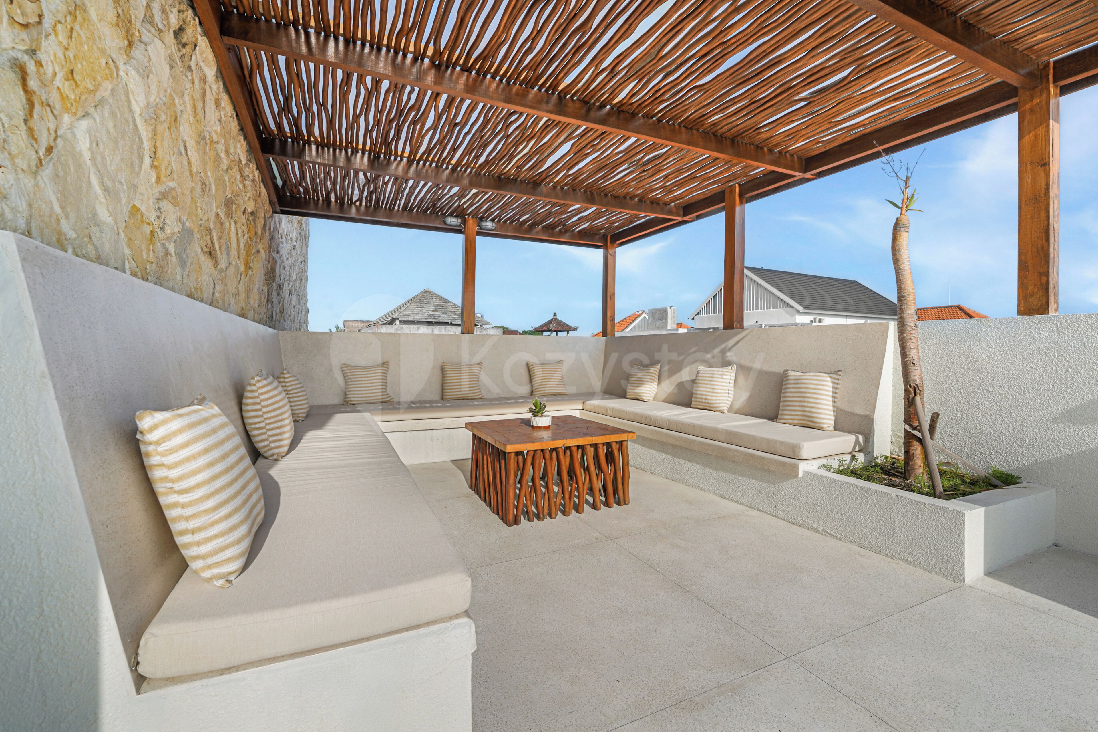 A covered outdoor seating area with a stone wall, a wooden pergola, and a view of buildings in the background.