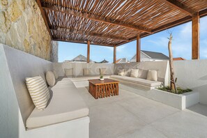 A covered outdoor seating area with a stone wall, a wooden pergola, and a view of buildings in the background. - Arkapura Villa by Kozystay - Canggu (Badung)