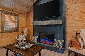 A cozy cabin interior with a fireplace, wooden walls, a television mounted above it, and a dining area with a table and chairs.