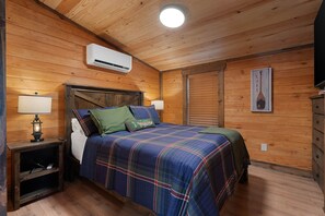Quarto de cabana de madeira com uma cama, duas luminárias de cabeceira, um criado-mudo e um ar condicionado fixado na parede.