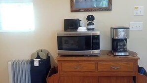 Una kitchenette con microondas, tostadora y cafetera sobre un mueble de madera, un radiador y un cuadro en la pared.