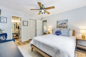 A hotel room with a bed, a desk, a chair, a ceiling fan, a bathroom with a mirror, and artwork on the walls. - Seahorse Stables - Spotless Coastal Cottage - 0.5 mile to Beach -  Sleeps 8 (Oak Island)