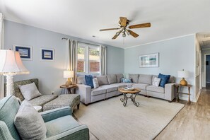 A living room with a sectional sofa, two armchairs, a coffee table, a ceiling fan, and a large window with curtains. - Seahorse Stables - Spotless Coastal Cottage - 0.5 mile to Beach -  Sleeps 8 (Oak Island)