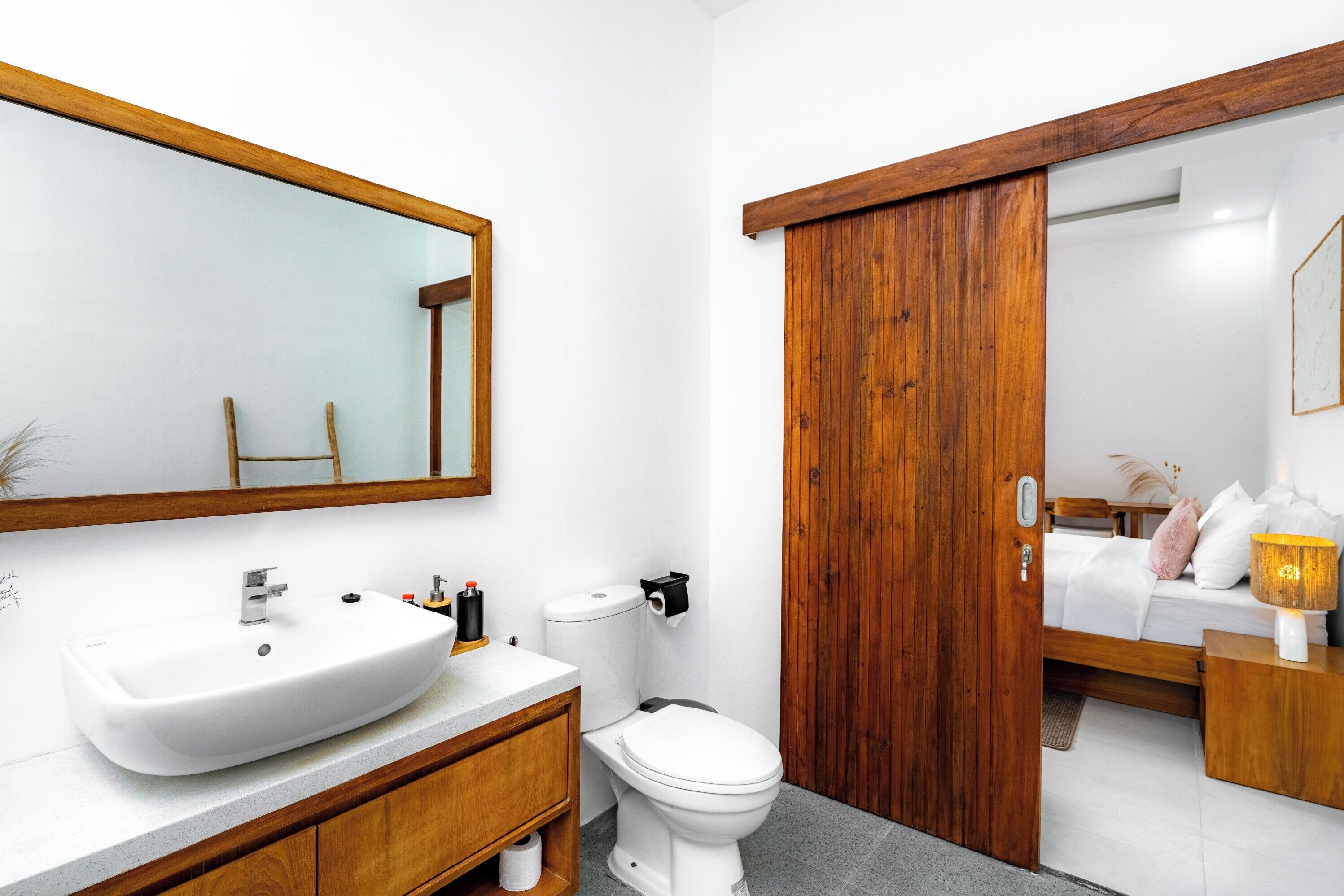 A modern bathroom with a large white sink, a toilet, and a wooden door leading to a bedroom.