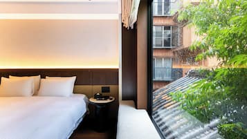 Habitación de hotel con cama, mesita de noche y vistas a un edificio y vegetación.