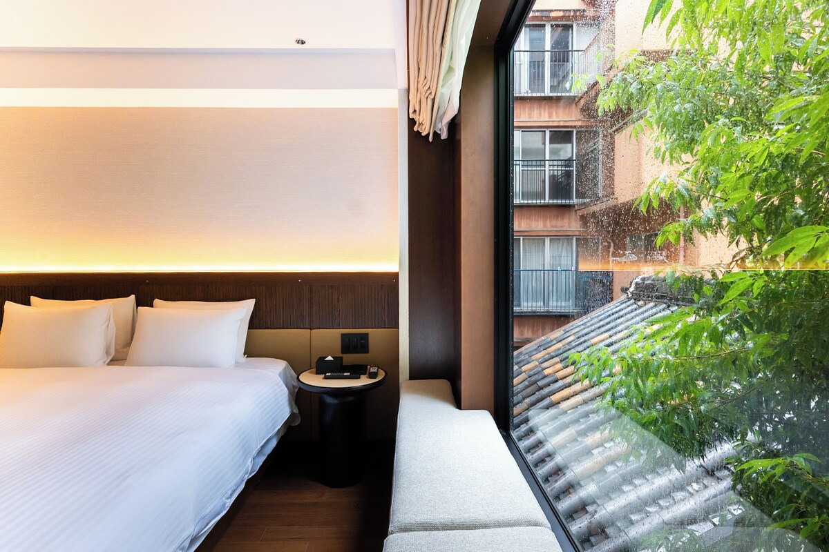 Habitación de hotel con cama, mesita de noche y vistas a un edificio y vegetación.