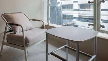 A room with a single chair and a table, a large window with a view of a building, and a plain wall.