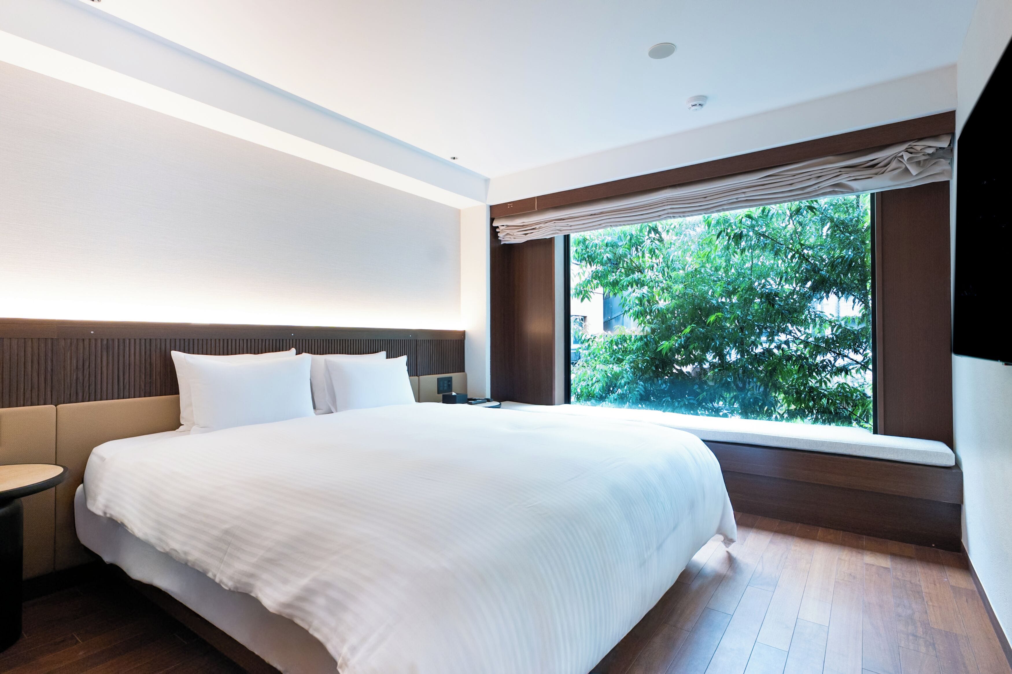 Une chambre d’hôtel moderne, dotée d’un grand lit, d’une table de chevet, d’une commode et d’une fenêtre donnant sur un paysage verdoyant.