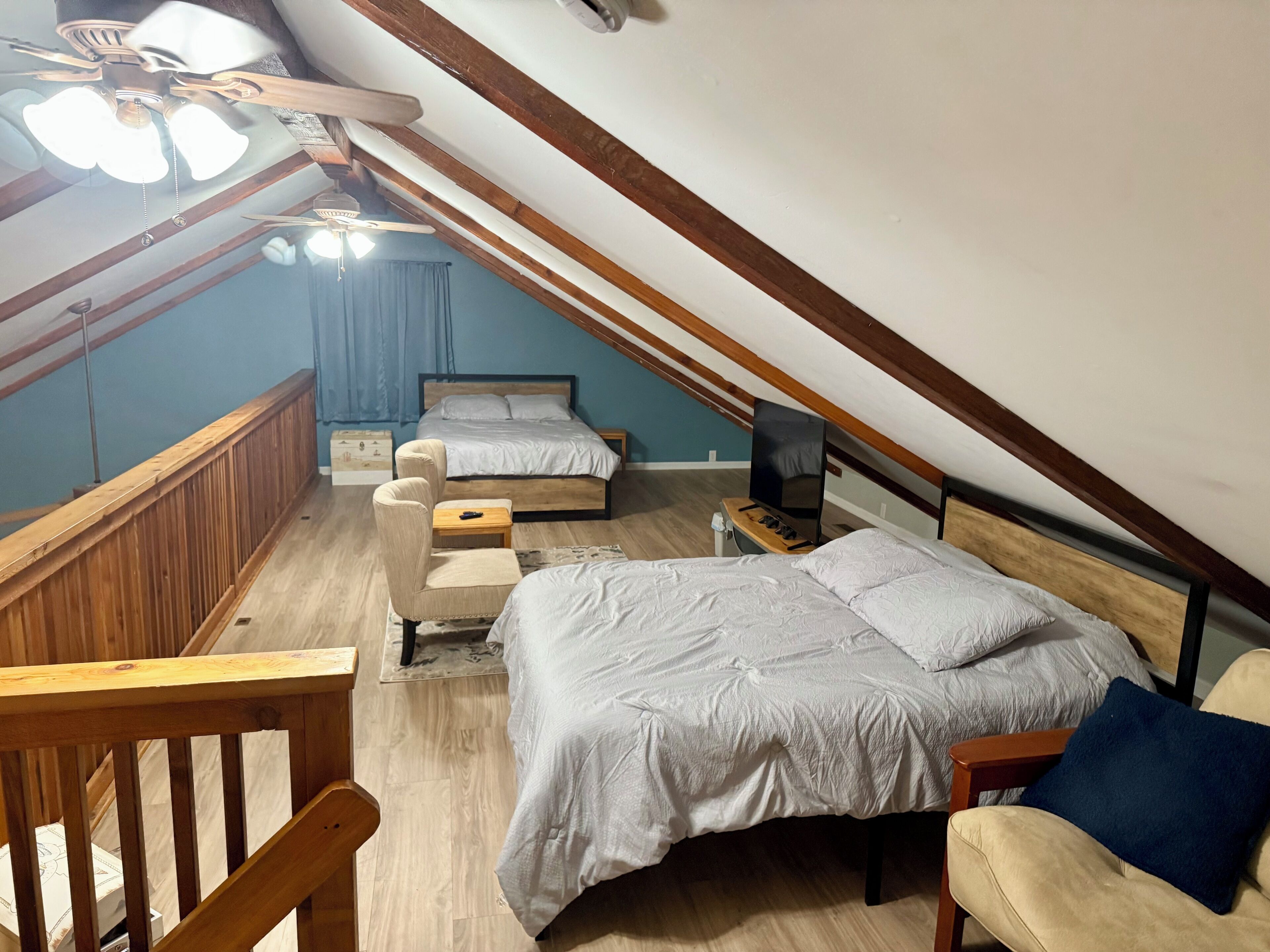 Une chambre avec un plafond en pente, un lit, un bureau, une chaise et un ventilateur de plafond.