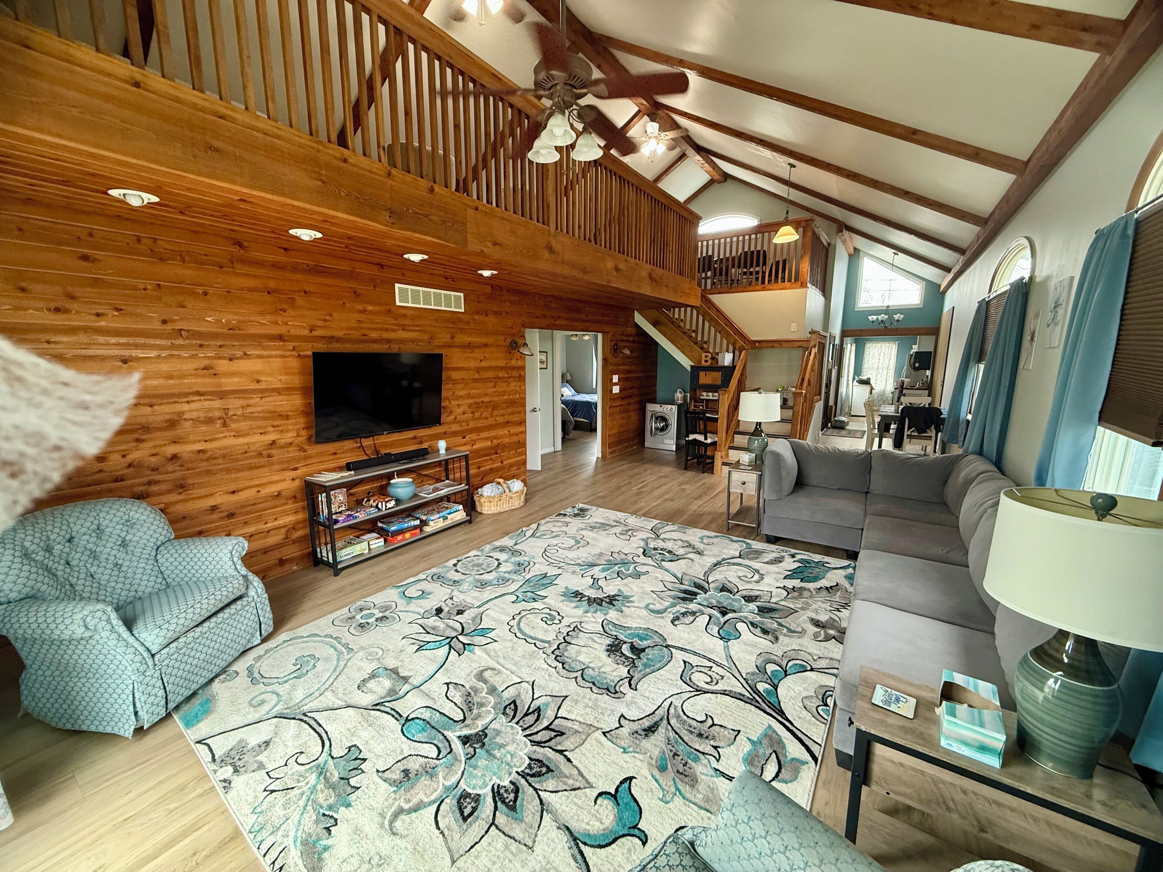 Een ruime woonkamer met een hoge plafond, houten balken, een grote flatscreen televisie, een bank, een gekleurd tapijt, en een trap naar een hoger gelegen verdieping.