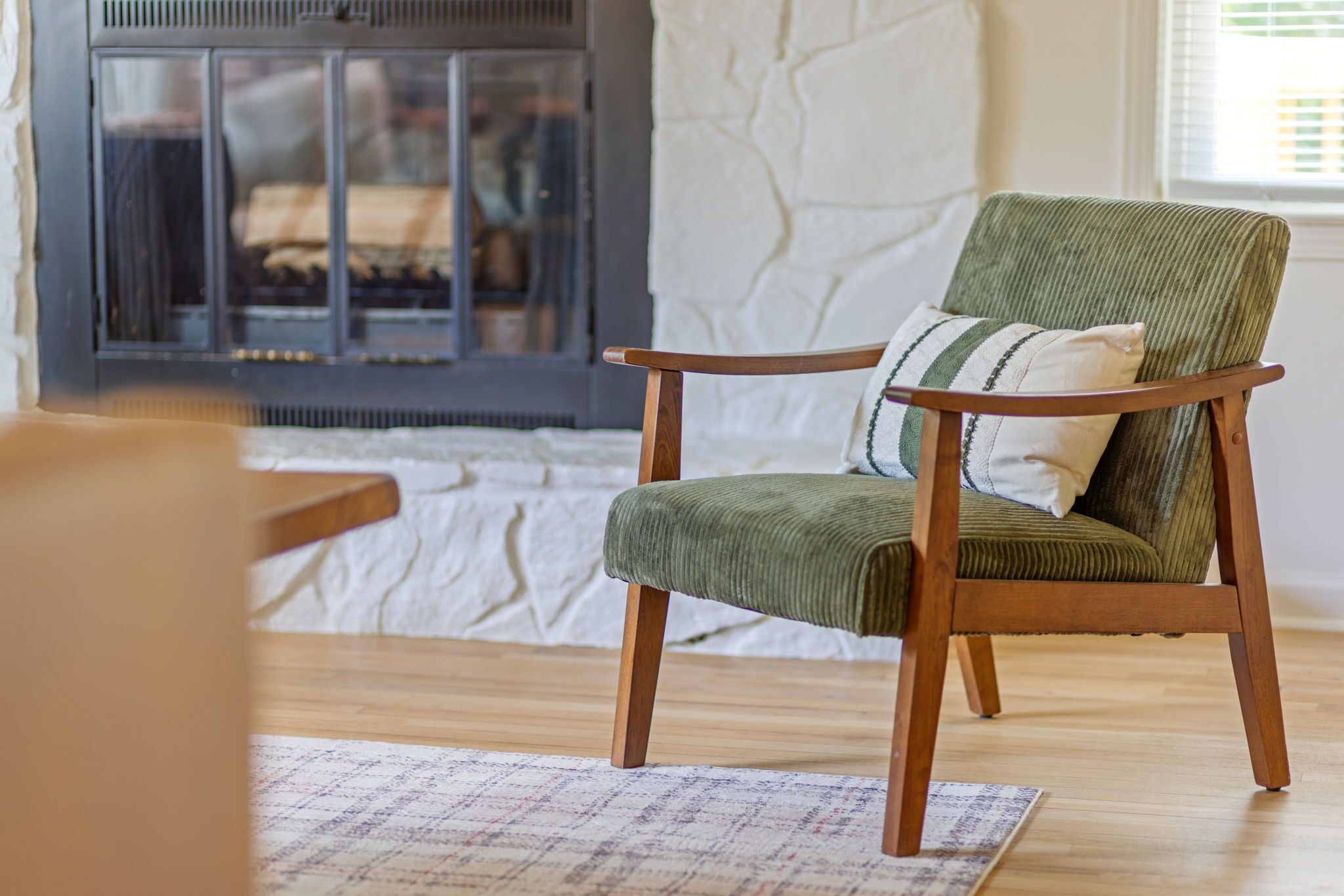 Un fauteuil vert recouvert de tissu, doté de pieds en bois et agrémenté d’un coussin à rayures, est placé devant une cheminée. Le mur derrière est blanc et présente une texture particulière.