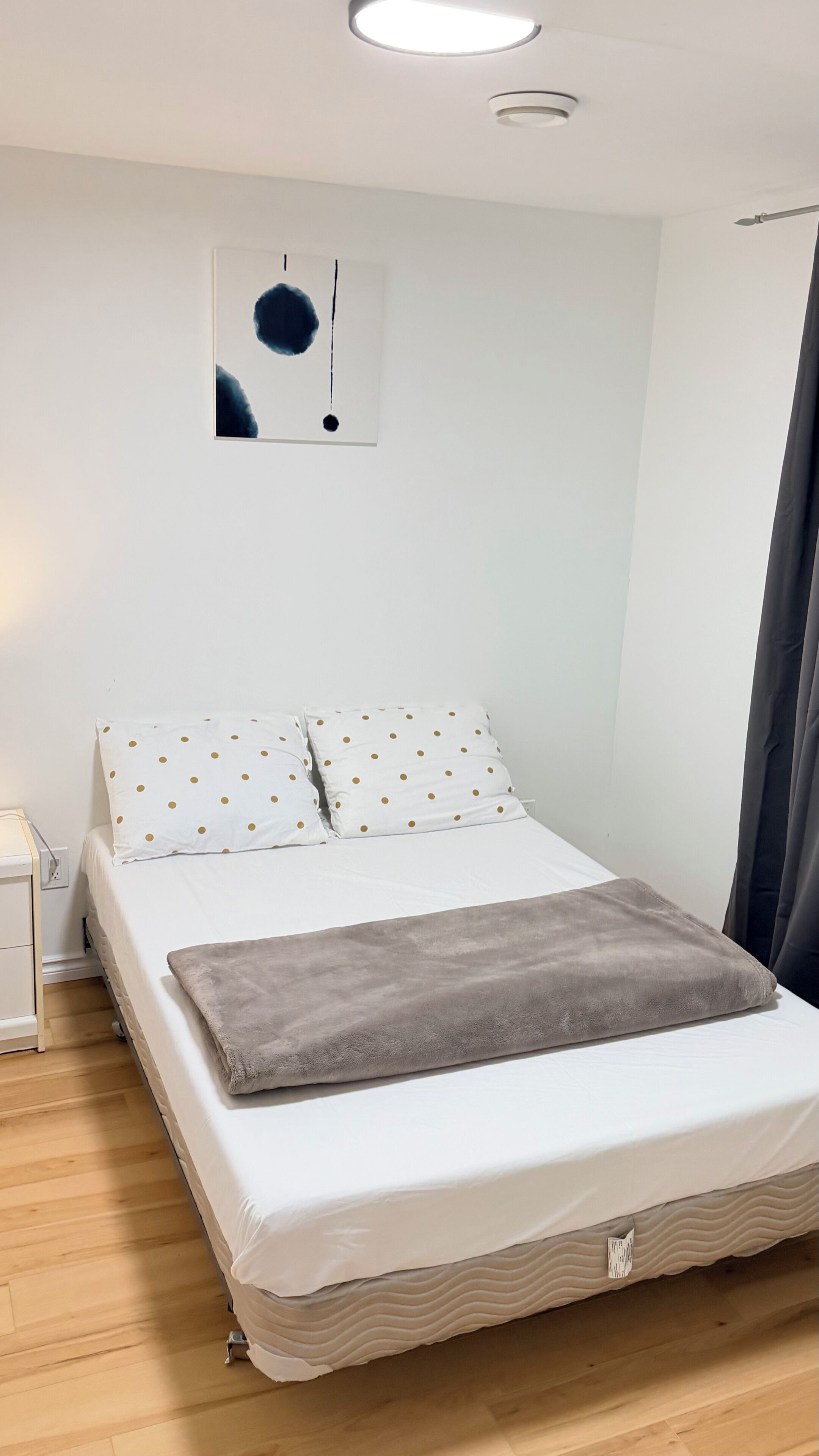 Une chambre à coucher avec un lit, du linge de lit blanc, un plaid gris, une table de chevet, une œuvre d’art accrochée au mur et une fenêtre avec un rideau.