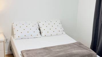 Une chambre à coucher avec un lit, du linge de lit blanc, un plaid gris, une table de chevet, une œuvre d’art accrochée au mur et une fenêtre avec un rideau.
