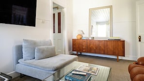 A living room with a sofa, a glass coffee table with magazines, a wooden cabinet with a mirror, and a door leading to another room. - Sisteron | Daylesford (Daylesford)