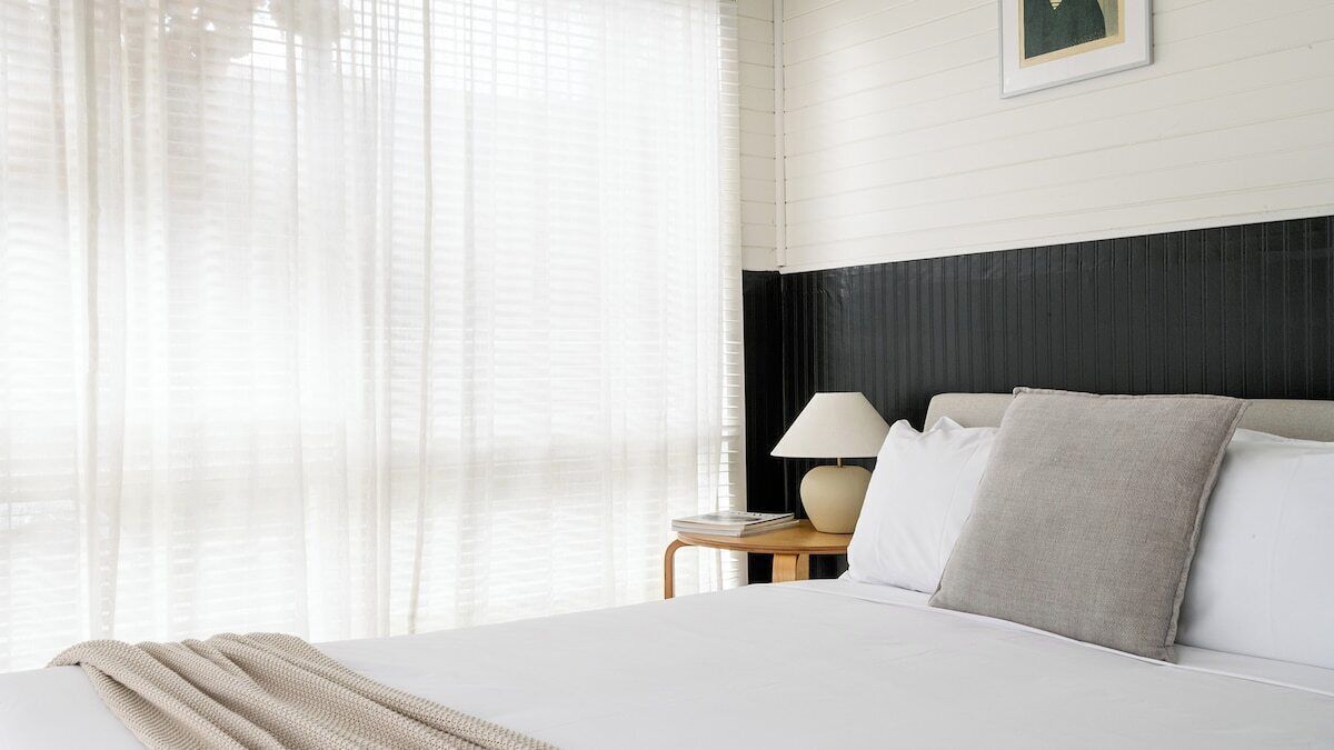A hotel room with a bed, a nightstand, a lamp, and sheer curtains.