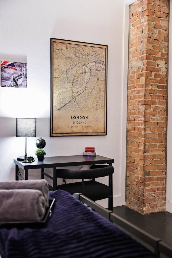 Een kamer met een kaart van Londen aan de muur, een klein tafeltje met een lamp, een model van een rode bus, en een bakstenen muur.
