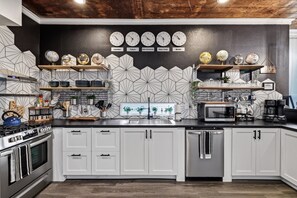 Een moderne keuken met witte kastjes, een gasfornuis, een magnetron en een koffieapparaat. Er hangen klokken aan de muur die verschillende steden aangeven.
