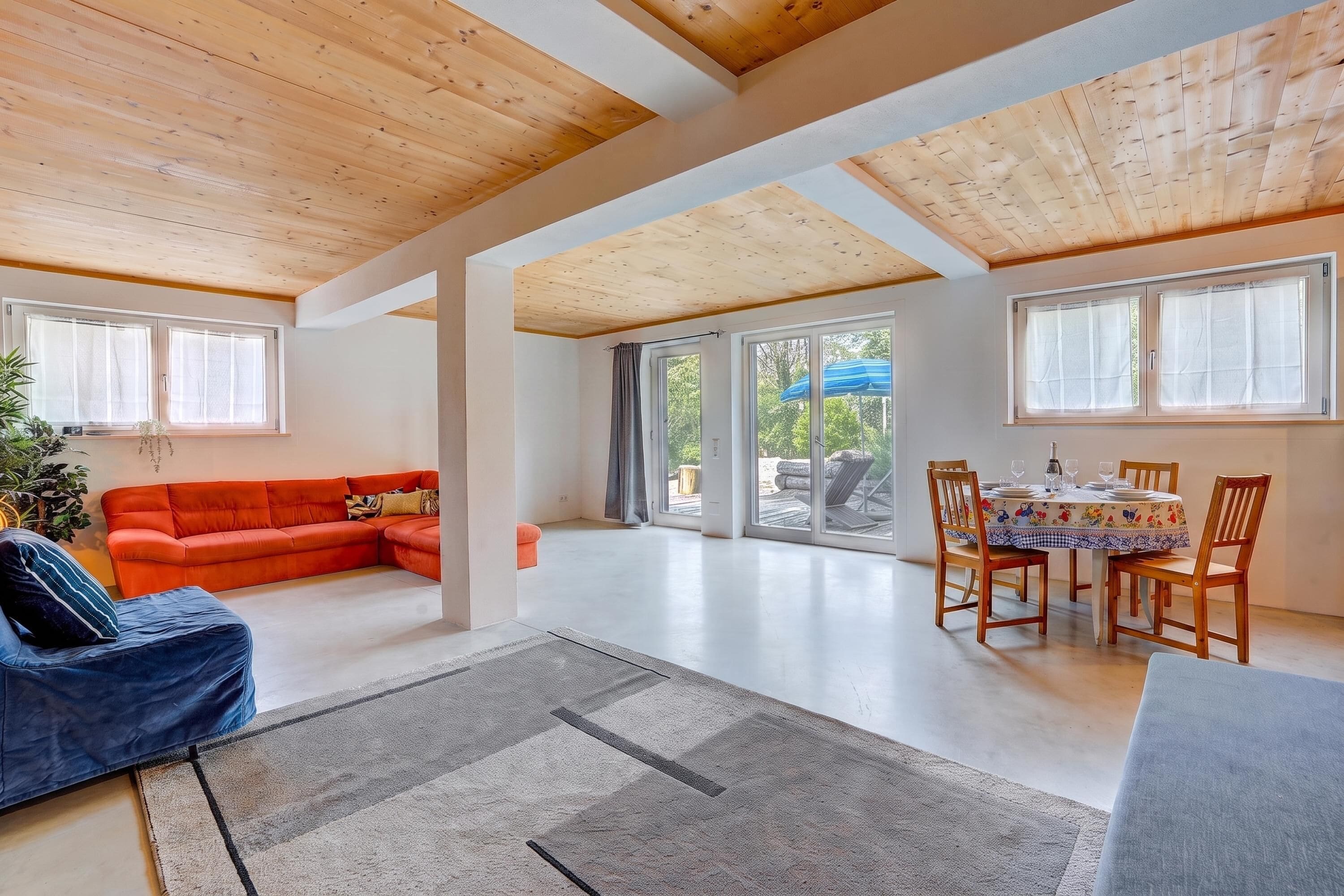 Amplia sala de estar con techo de madera, un sofá rojo, una mesa de comedor con sillas y ventanas grandes.