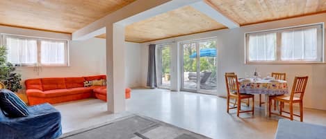 A spacious living area with a wooden ceiling, a red sofa, a dining table with chairs, and large windows.
