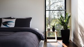 A bedroom with a bed, a nightstand, a potted plant, and a window with a view of trees.
