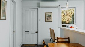 Ein Zimmer mit weißer Tür, einem gerahmten Bild an der Wand, einem Fenster mit Blick auf Bäume, einem Holztisch und einem Stuhl.