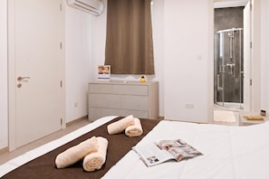 Ein Hotelzimmer mit Bett, Nachttisch mit Zeitschrift, kleiner Topfpflanze, Badezimmer mit Dusche und Fenster mit Vorhängen.