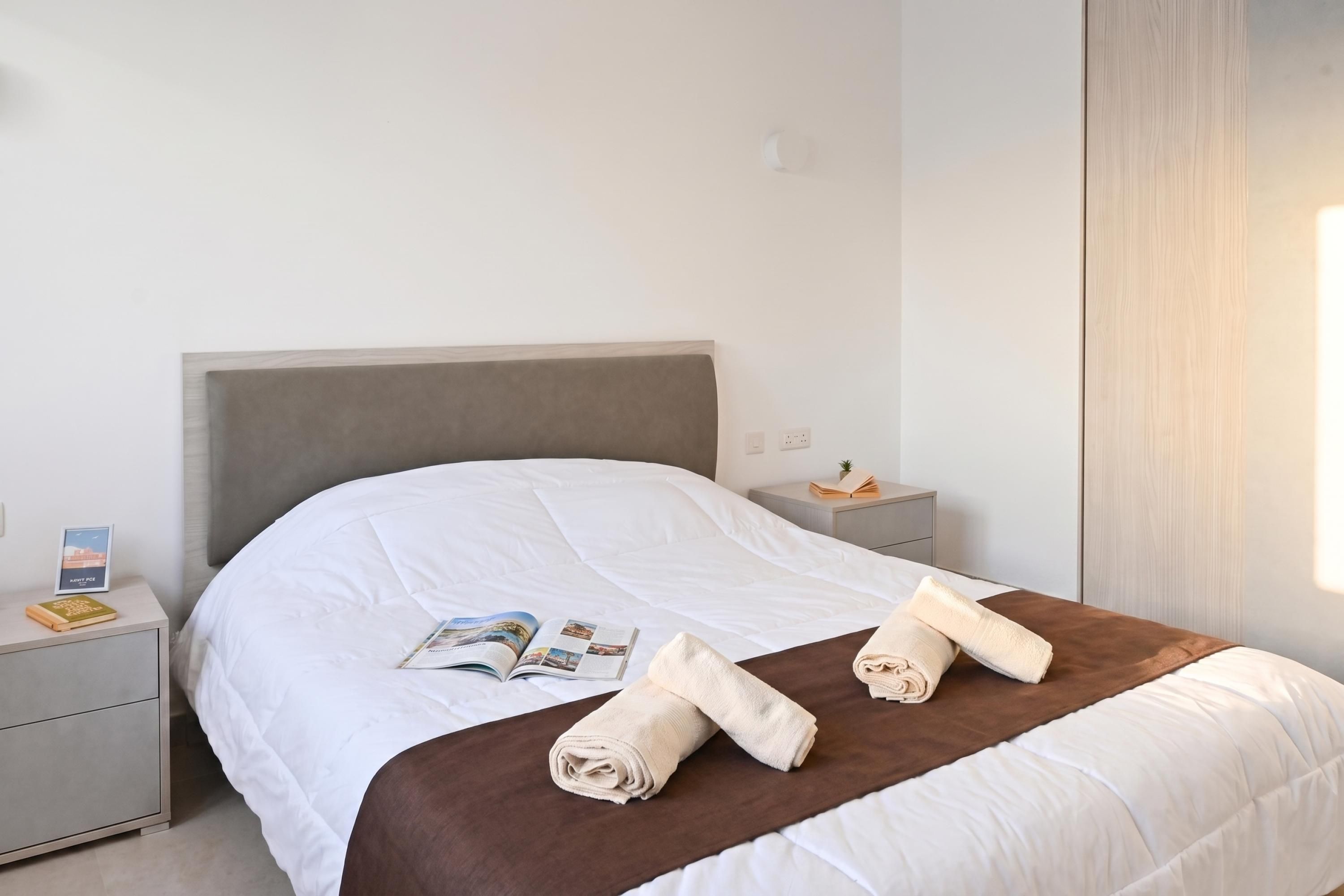 Una cama bien hecha con un detalle en marrón, una mesita de noche con un libro y una planta pequeña, y un interruptor de luz en la pared.