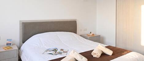 Una cama bien hecha con un detalle en marrón, una mesita de noche con un libro y una planta pequeña, y un interruptor de luz en la pared.