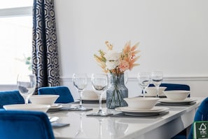 A hotel dining table set with plates, bowls, and wine glasses, a vase with flowers, and blue chairs. - Cosy Bungalow | Sleeps 7 | Free Hot Tub and WiFi (Leigh)