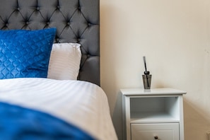 A neatly made bed with a tufted headboard, a white nightstand with a drawer, and a small table with a candle holder. - Cosy Bungalow | Sleeps 7 | Free Hot Tub and WiFi (Leigh)