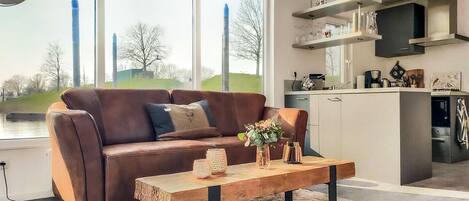 Un salon moderne avec un canapé en cuir marron, une table basse en bois et une vue sur une rivière et des arbres, visible à travers de grandes fenêtres.