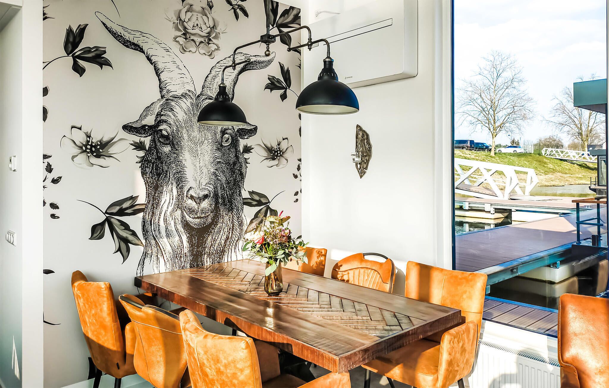 A dining area with a wooden table and orange chairs, a black and white floral wall mural, and a view of a bridge and greenery outside.