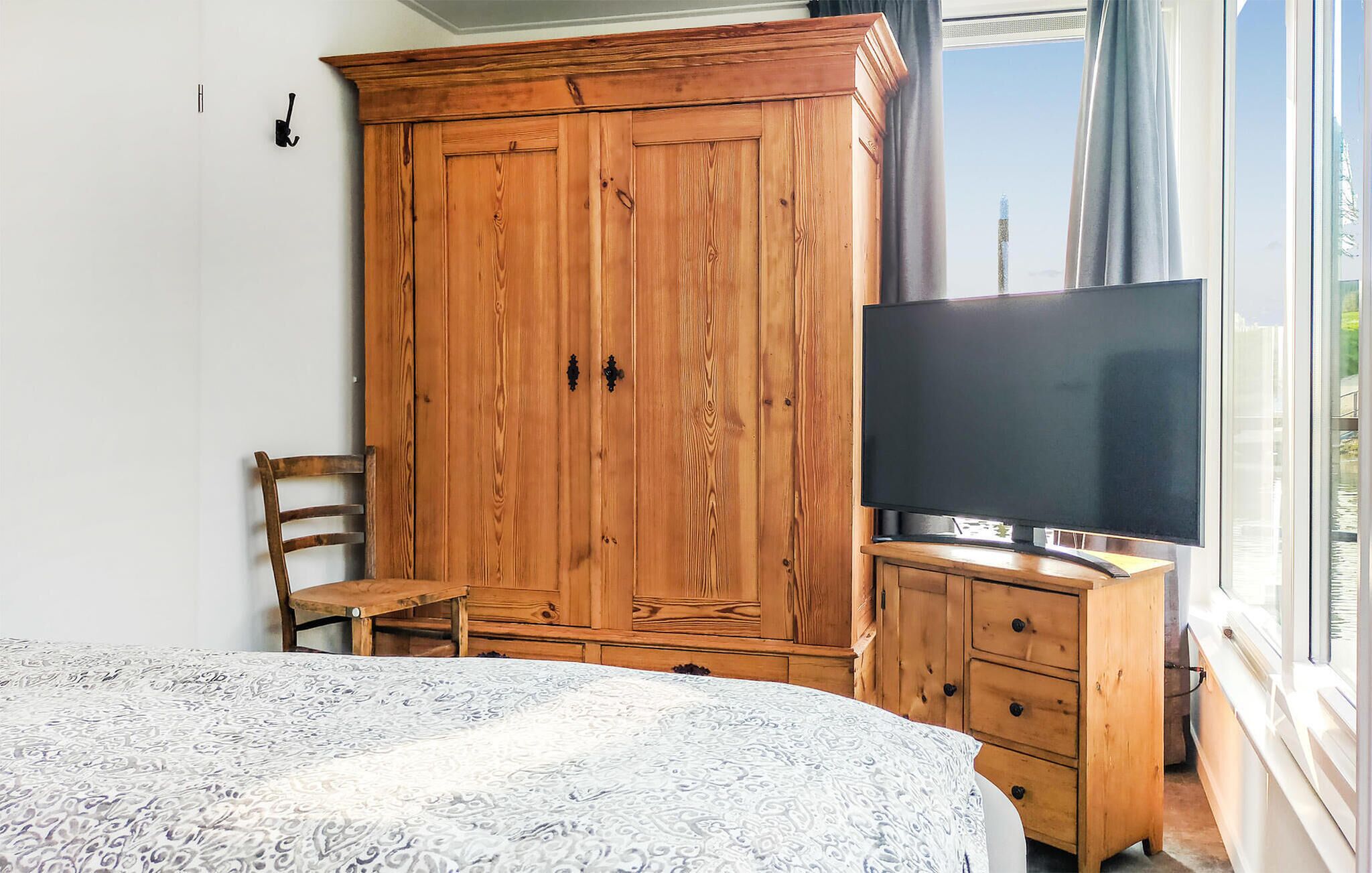 Un dormitorio con un armario grande, un televisor sobre un soporte de madera, una cama con colcha estampada y una ventana con vistas.