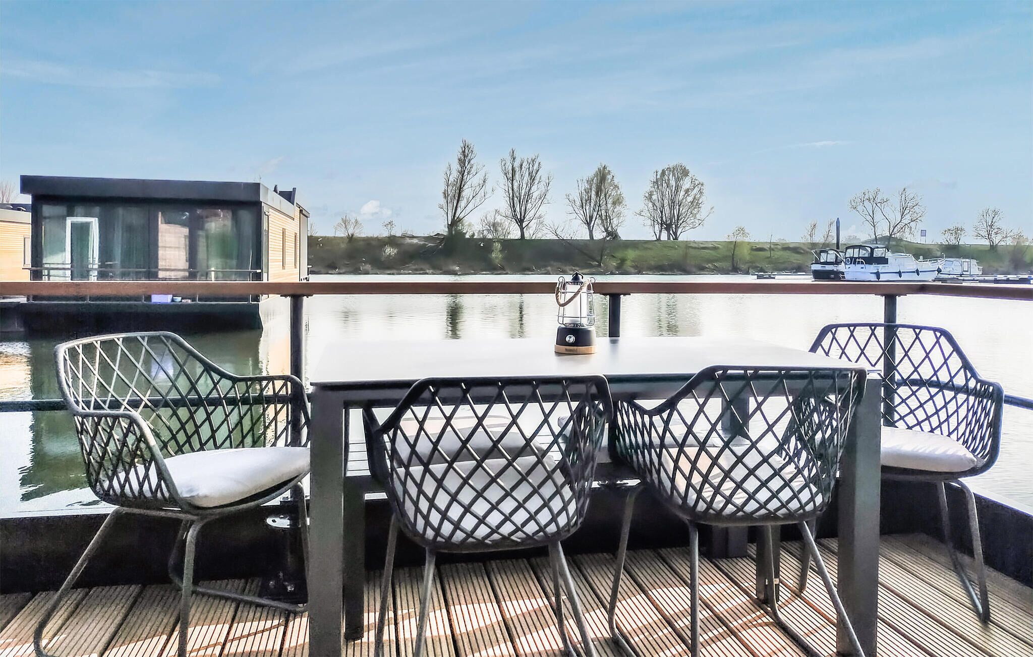 Eine Terrasse mit Tisch und Stühlen mit Blick auf ruhiges Wasser, im Hintergrund sind Boote zu sehen.