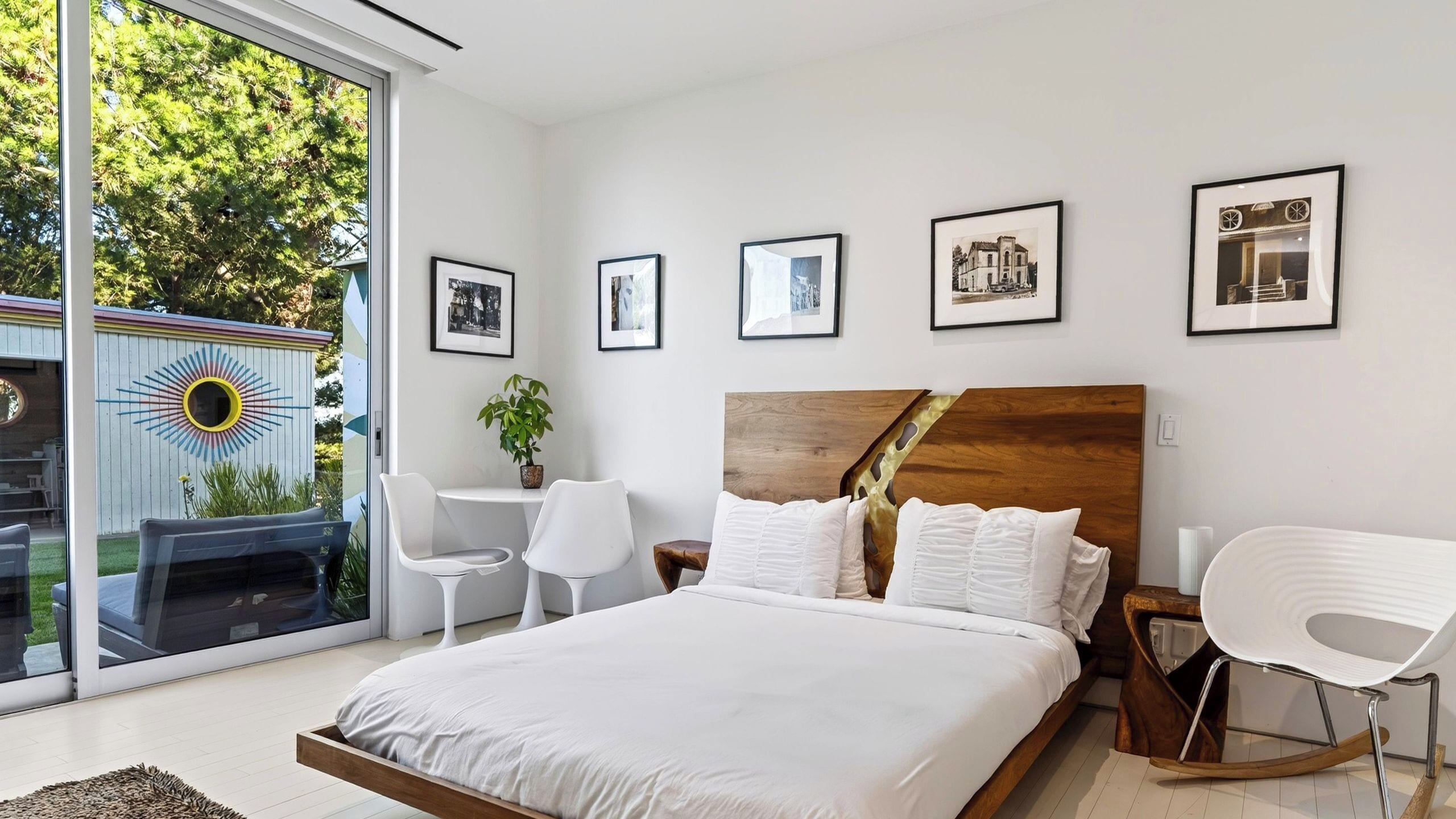 A modern bedroom with a large bed, a wooden headboard, white bedding, and a view of greenery through a sliding glass door.