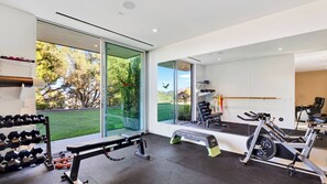 A modern gym with exercise equipment, a bench, and large windows offering a view of greenery and hills.