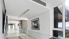 A modern hallway with glass railings, framed artwork on the walls, and large windows offering a view of the outdoors. - The Malibu Art Estate by Avantstay Infinity Pool + Theater + Game Room 20 Private Acres (Malibu)