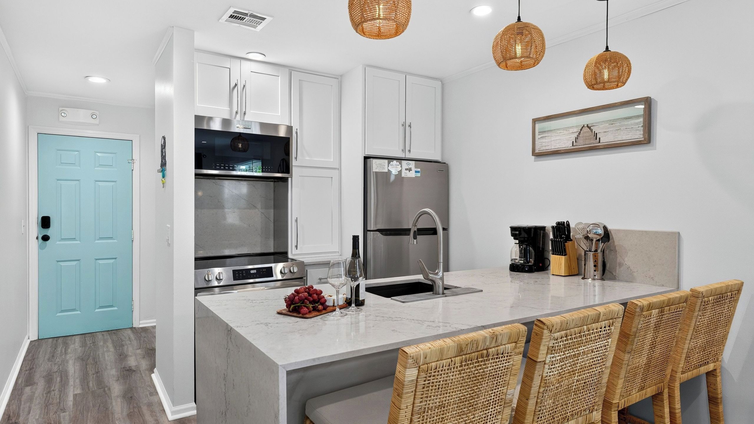 A modern kitchen with white cabinets, stainless steel appliances, a marble countertop, and wicker chairs.