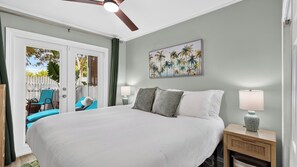 A bedroom with a large bed, a ceiling fan, a painting of palm trees, and a view of a patio with chairs and a fence. - Salty Palm Retreat by Avantstay Shared Pool + Private Porch Steps to Beach + Old Town (Key West)