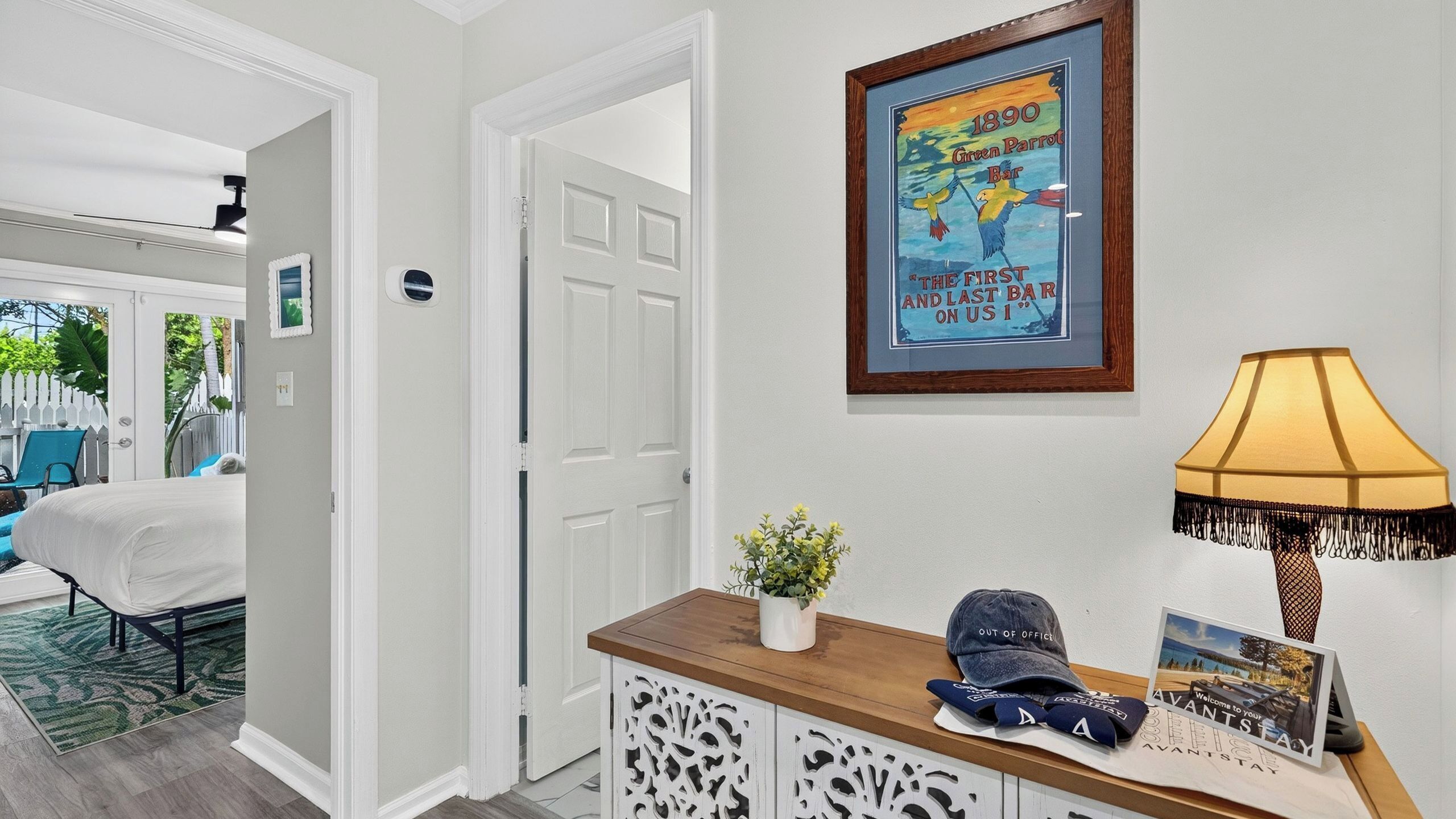 Un couloir d’hôtel avec une affiche encadrée, une lampe, un bureau avec une plante et un lit en arrière-plan.