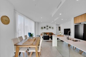 A modern kitchen with a dining area, a TV, and a clock on the wall. - Gorgeous 3rd Floor 3 BRM Apartment (Broadbeach)