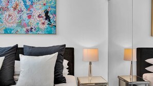 Une chambre d’hôtel avec un lit, deux tables de chevet, une lampe, un grand miroir et un tableau abstrait aux couleurs vives accroché au mur.