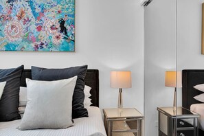 A hotel room with a bed, two bedside tables, a lamp, a large mirror, and a colorful abstract painting on the wall. - Gorgeous 3rd Floor 3 BRM Apartment (Broadbeach)