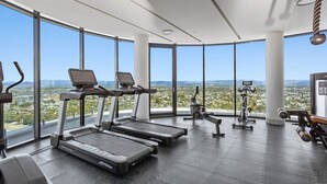 Une salle de sport équipée de tapis de course, de vélos d’appartement et de poids, avec de grandes fenêtres offrant une vue sur la ville.