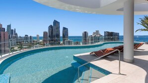 Une piscine à débordement située sur le toit, offrant une vue panoramique sur la ville, avec des chaises longues et un palmier.