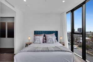 Ein modernes Schlafzimmer mit einem großen Bett, zwei Nachttischen und Blick auf die Stadt durch große Fenster.