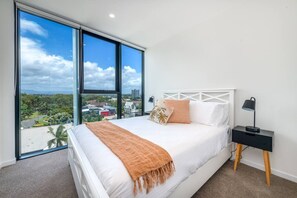Une chambre avec un grand lit, une table de chevet, une lampe et une vue sur le paysage urbain grâce à de grandes fenêtres du sol au plafond.